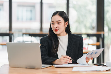 Professional woman working on laptop, analyzing data with documents in hand. She appears focused and engaged in her task, showcasing modern office setting