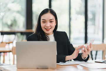 professional woman in black blazer smiles while using smartphone and laptop in modern office setting, showcasing productivity and engagement