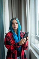 Vertical portrait of beautiful blue haired hipster caucasian woman standing in front of window taking selfie with her mobile phone. Looking at camera. Using technology.