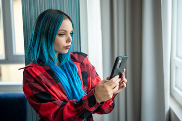 Portrait of beautiful blue haired caucasian hipster woman standing in front of window taking selfie with her mobile phone. Using technology.