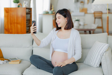 Young Asian happy pregnant woman is sitting and using mobile phone on sofa and touching her belly. Family plan, pregnancy, motherhood, people and expectation concept