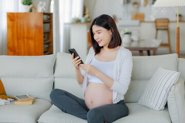 Young Asian happy pregnant woman is sitting and using mobile phone on sofa and touching her belly. Family plan, pregnancy, motherhood, people and expectation concept