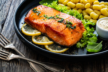 Seared salmon steak with gnocchi and lemon slices served on black plate on wooden table
