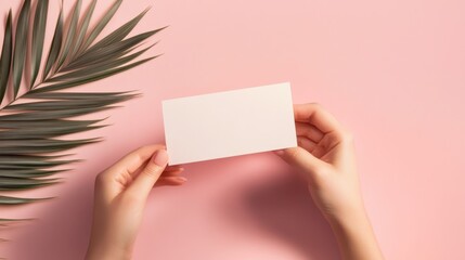 Obraz premium A person holds a blank card against a pink background, complemented by a green palm leaf, creating a minimalist and stylish aesthetic.