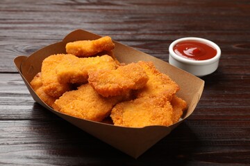 Tasty chicken nuggets in paper container and sauce on wooden table, closeup
