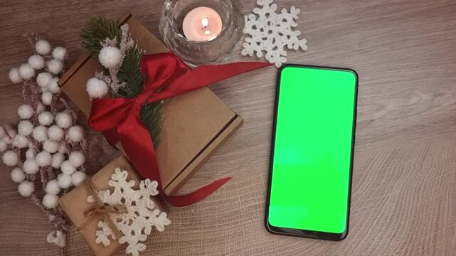 Green screen smartphone lying vertically on wooden table next to burning candle, gifts fnd DIY Christmas decorations. Christmas mockup top view.
