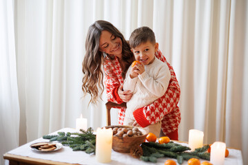 Smiling woman hugging cheerful son eating tangerine near decorated dinning table for Christmas...