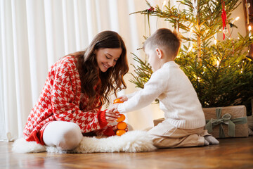 Fototapeta premium Boy playing with tangerines near smiling mother and Christmas tree at home