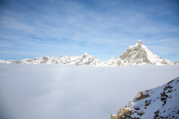 A stunning view of snow-covered peaks, with the iconic Matterhorn rising above a sea of clouds under a clear blue sky. The image captures the majestic beauty of the Swiss Alps in winter