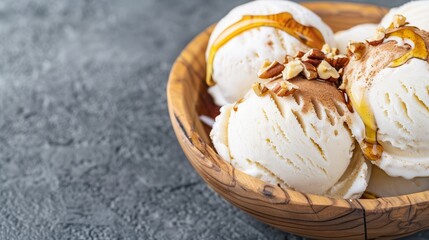 A creamy scoop of vanilla ice cream topped with caramel and chopped nuts in a wooden bowl, set against a textured gray background.