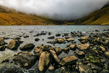 Rocks litter the shore of a lake with a layer of fog blanketing the hills surrounding it.