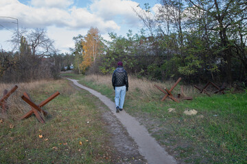 A solitary figure strolls down a narrow dirt path in a neglected area, flanked by rusty barricades...