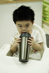 Asian boy drinking water in silver glass, sitting on high chair at home