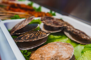 A detailed view of scallop shells on fresh lettuce with skewers of grilled meat in the background, depicting a Shanghai market setting.
