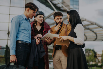 A group of young professionals are engaged in a business discussion outside a contemporary office building, reviewing documents and strategies. The diverse team appears focused and collaborative