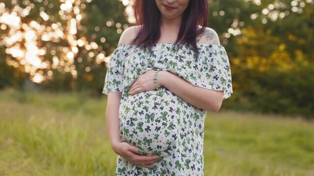 Pregnant caucasian woman in summer dress posing outdoors. Close up of young lady holding two hands on her pregnant belly while standing at green garden. Relaxation on nature during summer days