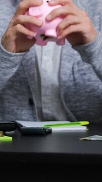 Vertical video. Man sits at his office desk, eagerly emptying a pink piggy bank in the office setting, coins scattering across the surface, savings, budgeting and financial planning concept