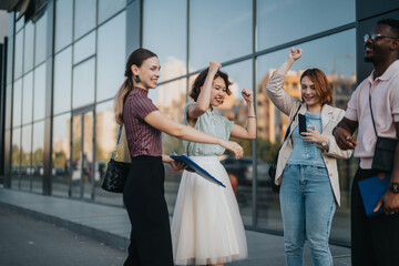 A multicultural group of business professionals cheerfully celebrating an achievement outside a modern urban building, showcasing teamwork and success in a diverse corporate environment.