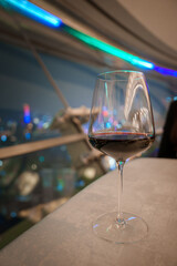 A wine glass filled with red wine sits on a table, with a blurred view of Shanghai's illuminated cityscape and modern architecture in the background.