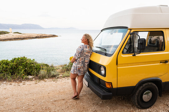 Young blonde woman enjoying the sea view in her yellow retro camper van parked on a dirt road above the sea on Korcula island, Croatia