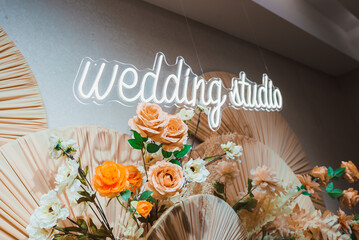 A neon sign reading 'wedding studio' is surrounded by artificial flowers in peach, orange, and...