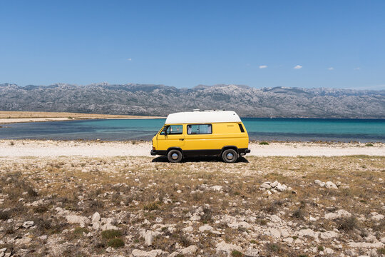 Yellow retro camper van parked on the rocky wild beach on Pag island with the mountains in the background, Croatia