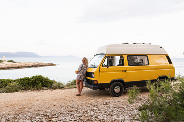 Young blonde woman enjoying the sea view in her yellow retro camper van parked on a dirt road above the sea on Korcula island, Croatia