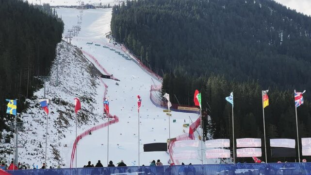 International flags wave above a bustling ski slope in Bansko, showcasing a vibrant winter sports scene against a backdrop of snow-covered mountains and forest.