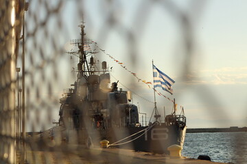 Guarded Strength: Warship and Greek Flag Through the Fence © Dakota B. Oldeman