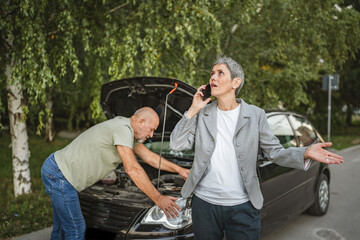 Senior man check car engine while mature woman calls for assistance