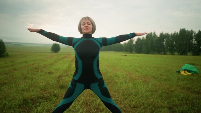 Woman in green and black suit on yoga mat practicing triangle pose with arms and legs extended, standing in an open grassy field, surrounded by greenery and trees under a cloudy sky