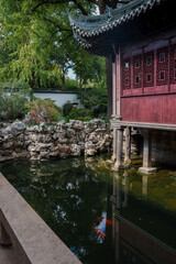 Naklejka premium A red wooden pavilion with lattice windows stands over a pond in a Shanghai garden, surrounded by rocks and greenery, reflecting traditional architecture.