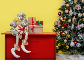 Vintage Harlequin doll on red dresser with gift in hands near Christmas tree on yellow background...