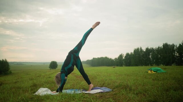 Woman in green and black suit practicing standing split yoga pose outdoors on a yoga mat in a grassy field under a cloudy sky, stretching her body downward as part of a wellness routine