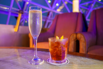 A tall flute of champagne and a cocktail with citrus garnish sit on a table. Plush armchairs and a...