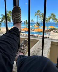 Woman wearing tennis shoes propping her feet up on the rails of the balcony of hotel room overlooking gorgeous view of beach and ocean on Oahu Honolulu HI.