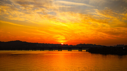 A breathtaking sunset paints the sky in fiery hues of orange and yellow. The sun dips below the horizon, casting a golden glow on the calm river and silhouetting the bridge.