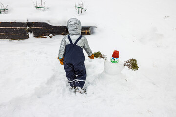 Child in winter clothing stands next to small snowman decorated with red pot hat and evergreen branch arms. Sweden.