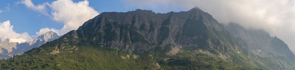 Panoramic view of majestic mountain range partially covered in clouds with rugged peaks and dense greenery, blue sky, forest, alpine landscape. Tonale Pass, Italy. Summer vacation. Dolomite Alps