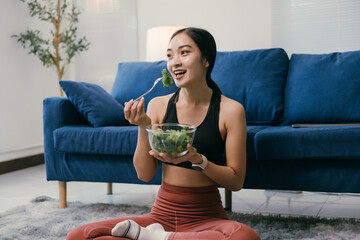 Happy athletic woman eating healthy salad after workout, sitting on floor in living room, enjoying her meal while relaxing on carpet in front of sofa