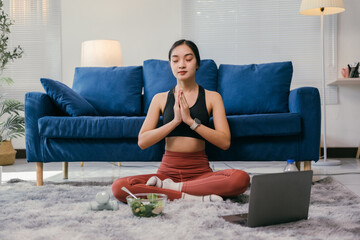Healthy asian woman practicing yoga and meditating at home on a carpet in her living room after a fitness workout, with a bowl of salad and a bottle of water nearby, enjoying a healthy lifestyle