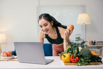 Asian nutritionist holding an apple and smiling while giving online consultation about healthy eating and dieting through video call using laptop in her modern and cozy home office