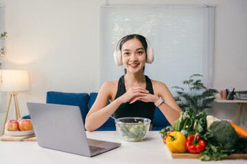Young nutritionist wearing headphones and sportswear is smiling while sitting at a table with a...