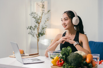 Happy asian nutritionist wearing headphones and using laptop while following online healthy eating...