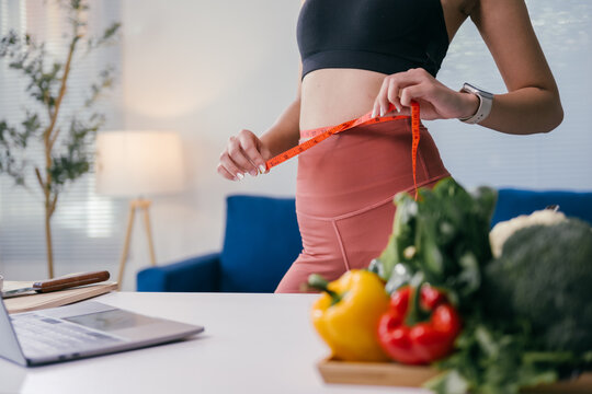 Young woman in sportswear measuring her waistline with a tape measure while standing near a table with fresh vegetables and a laptop, promoting healthy lifestyle and weight management