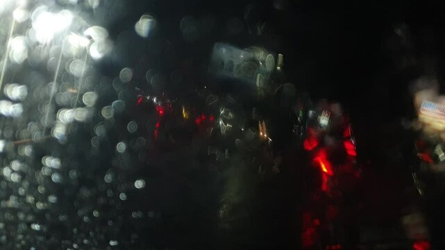 Rain at night on the side of the highway seen from the perspective of inside the car, you can see raindrops sticking to the car window. There is light from street lights, cars and other vehicles passi
