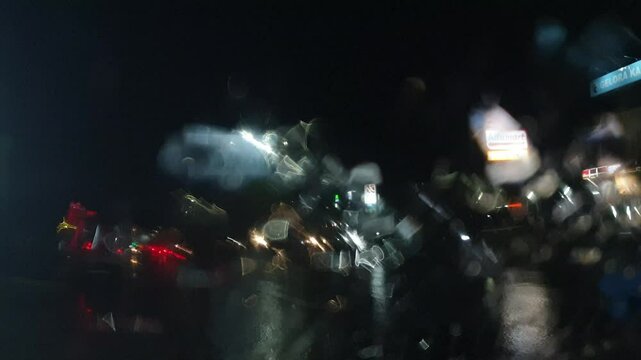 Rain at night on the side of the highway seen from the perspective of inside the car, you can see raindrops sticking to the car window. There is light from street lights, cars and other vehicles passi