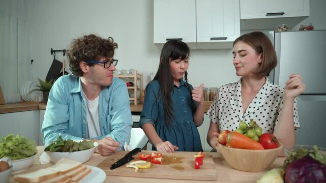 Attractive caucasian father and mother spend time together to cook fresh meal with asian daughter. Dad and mom preparing fresh vegetable for making salad at kitchen and eating vegetable. Pedagogy.