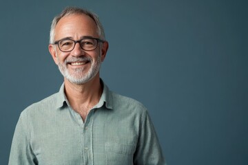 Fototapeta premium Senior male presenter in a casual shirt, smiling, standing against a solid blue-gray background with space for text