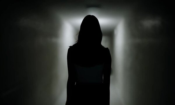 Dark exposure silhouette of a woman view from the back in defocus. A female figure is walking along a dark corridor, moving away from the camera